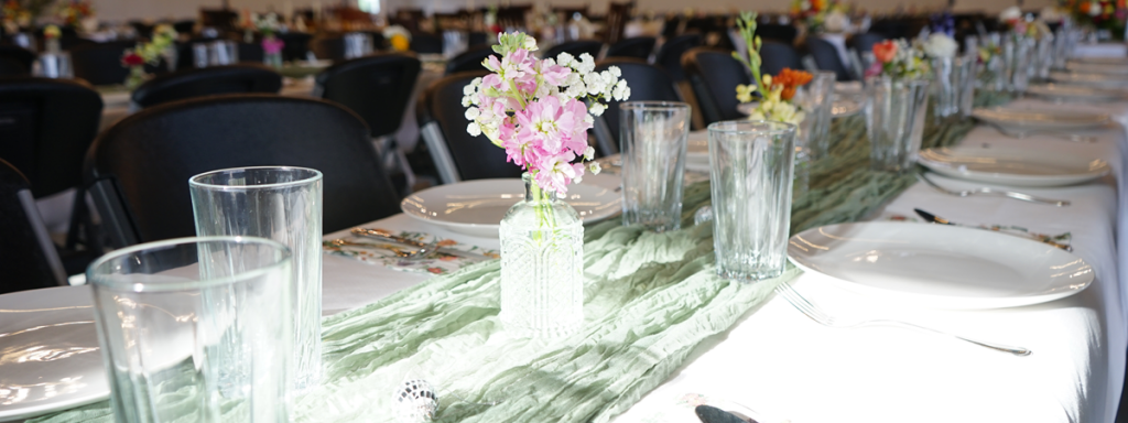 A long banquet table is set with white plates, clear glasses, and silverware. The table is decorated with a green cloth runner and small vases of pink and white flowers. Black chairs are arranged along both sides.