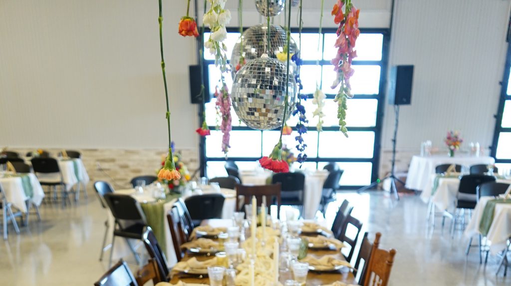 A decorated event space with tables set for a meal, disco balls and colorful flowers hanging above the center, and black chairs arranged around the tables.