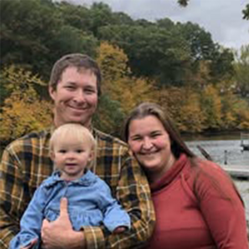 A smiling couple stands outdoors, with the man holding a young child. They are in front of a river with a backdrop of trees displaying autumn foliage. The child is wearing a blue outfit, and the adults are dressed in casual fall attire.