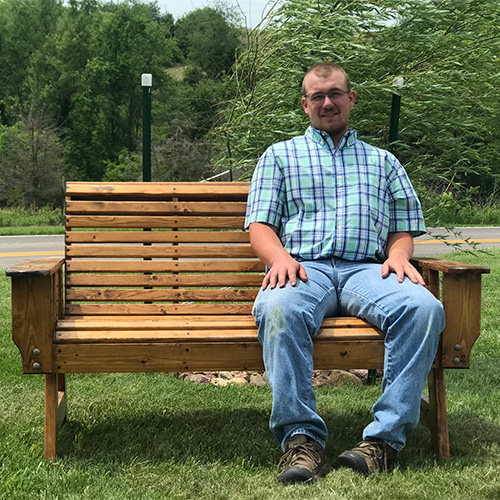 Andrew Anderson, in a plaid shirt and jeans, sits on a wooden bench in a grassy area with trees gently swaying in the background. It's a sunny day.