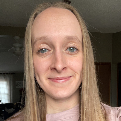 Kayla Clausen, with her long straight hair, is smiling slightly at the camera in a pink top. The background features a dimly lit interior with a ceiling fan and curtains.