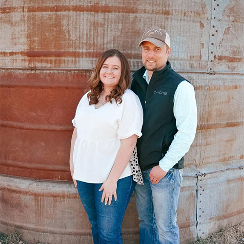 Kaylee Churchill and a man stand together in front of a large, rust-colored metal tank. She wears a white blouse and jeans, while he sports a cap, black vest, light shirt, and jeans. They both smile warmly at the camera.