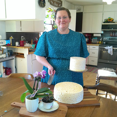 Roberta Kauffman, in a teal dress, stands in the kitchen holding a knife and a round cheese. On the table, two large cheese wheels rest on a wooden board beside a small potted plant with vibrant purple flowers.