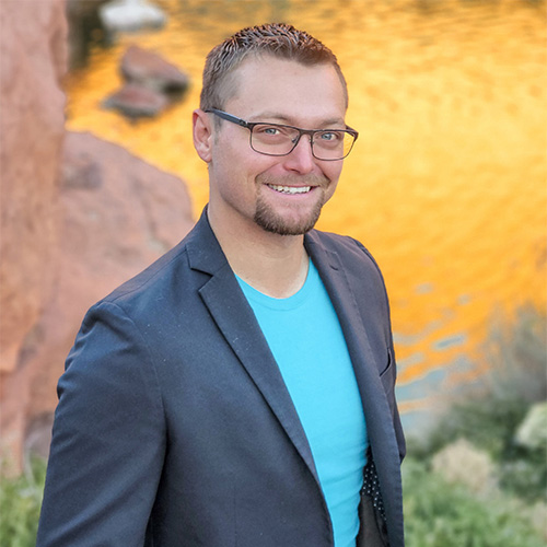 Jamon Mysliwiec wearing glasses, a blue T-shirt, and a dark blazer stands outdoors. Behind him, there are rocks and a body of water reflecting golden sunlight. He is smiling and looking at the camera.