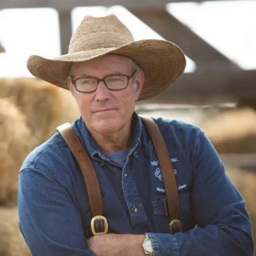 Joel Salatin wearing a straw cowboy hat, glasses, and a blue denim shirt, stands in front of hay bales. He has leather suspenders and appears to be outdoors, with a part of a wooden structure visible in the background.