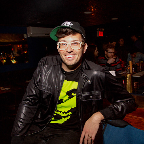 Luke Thayer, sporting a black leather jacket, graphic tee, cap, and glasses, is smiling at a dimly-lit bar with tables and patrons in the background.