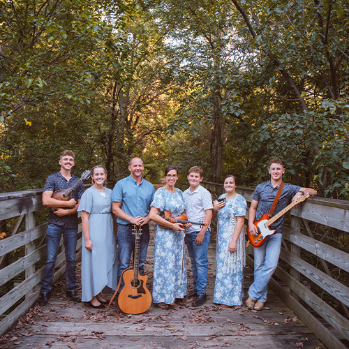 A group of seven, known as The Peachey Family Band, stands on a wooden bridge enveloped by trees. They hold musical instruments—guitars, a violin, and a tambourine—creating a harmonious scene in the forest's daylight.