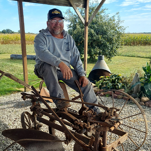 David Savage sits on an old rusted piece of farm equipment under a wooden shelter, sporting a cap and gray hoodie. Behind him stretch fields, with a clay chiminea and some plants adding rustic charm. Above, the sky is partly cloudy.