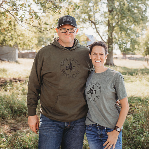 A man and woman, with bright smiles, pose outdoors under the trees. The man sports a dark hoodie and cap, while Kim Lauffer stands beside him in a gray T-shirt. Behind them, the grassy area and fence add to the serene backdrop.