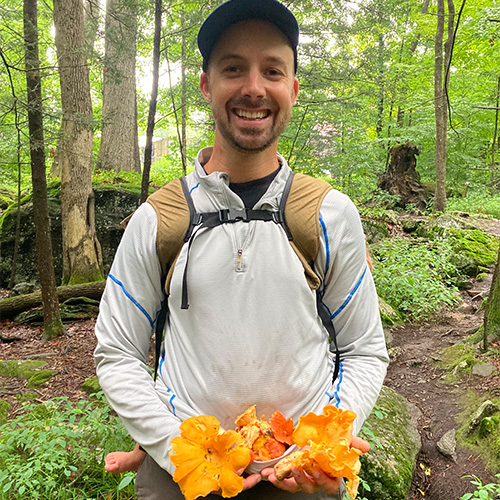 Man wearing a cap and backpack smiles while holding a handful of bright orange mushrooms in a forest setting with green trees and foliage.
