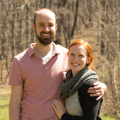 A smiling couple stands outside in a wooded area. The man, Peter Kruger, wearing a pink shirt, has his arm around the woman with red hair, who is wearing a gray scarf and black sweater. The background shows bare trees and grass, suggesting early spring or fall.