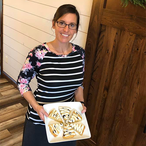 Tina Peachey with glasses and a floral and striped top holds a white tray with six triangular pastries topped with icing. She is standing in a room with wooden flooring and a wooden door in the background.