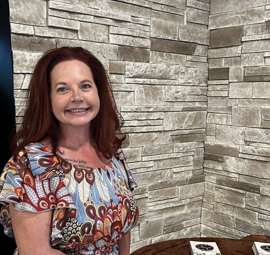Ashley with long red hair and a colorful patterned dress smiles while standing in front of a textured stone wall. A table with a closed book is partially visible in the foreground.